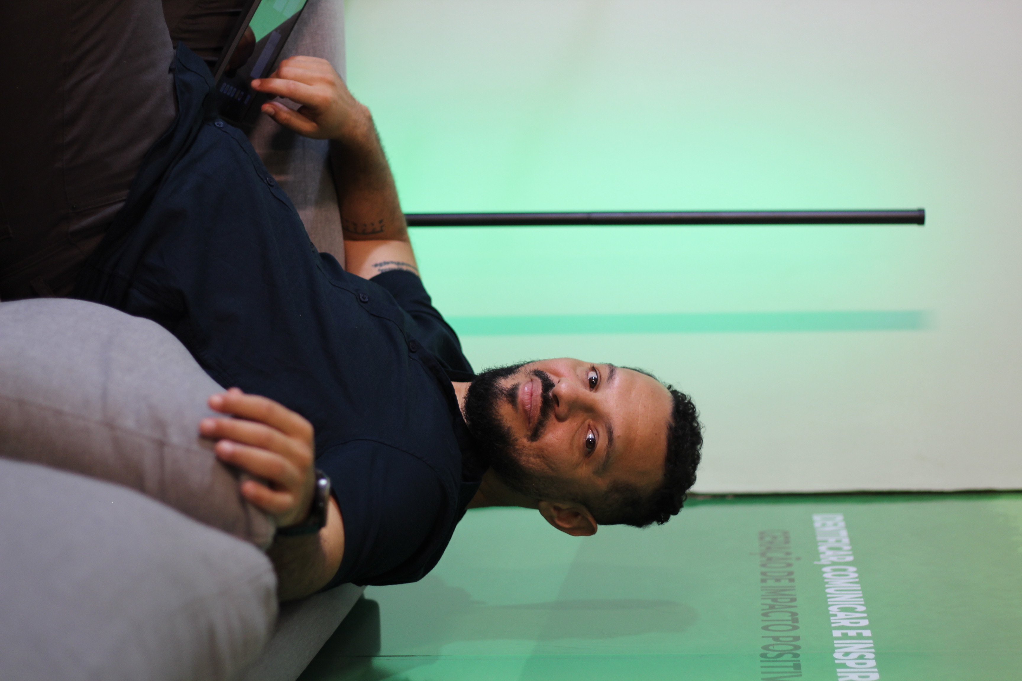 Adriano Diamarante sentado em um sofá, sorrindo, com parede verde ao fundo.